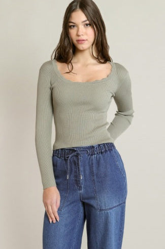 A woman modeling a ribbed long-sleeve scoop-neck top in sage green, paired with blue jeans, against a plain light beige background.