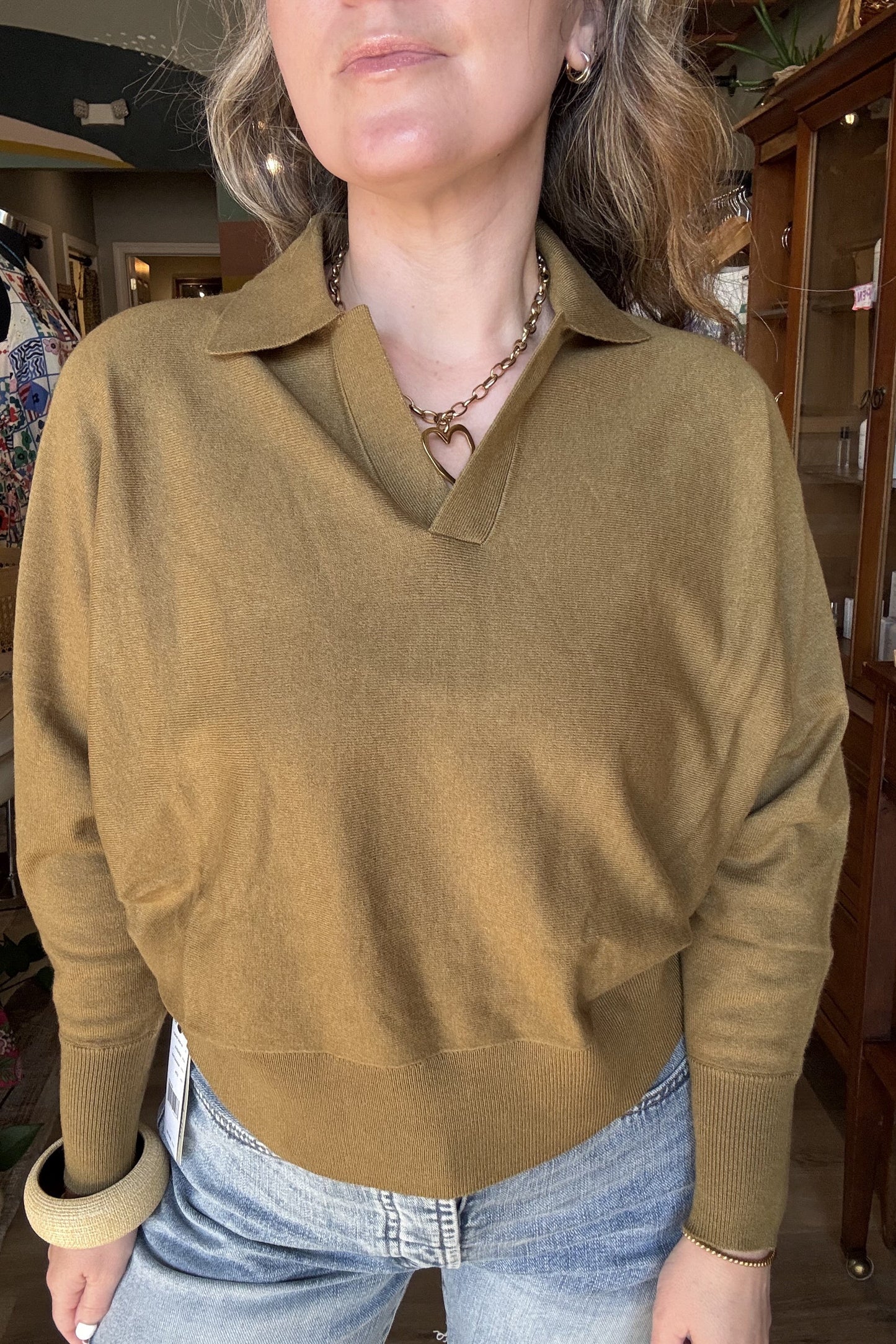 A woman wearing a camel-colored knit polo sweater with a collar and long sleeves, paired with light blue jeans, standing inside a retail store.