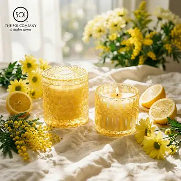 Two yellow candles in textured glass holders with flowers and lemons on a light surface, featuring 'The Soi Company' logo.