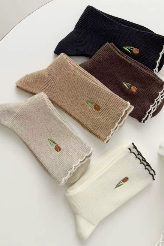 Four pairs of ribbed ankle socks arranged on a white surface, each with a small embroidered tulip near the top. The socks come in black, dark brown, taupe beige, and off-white/cream, and two have black lace trim while the others have white lace trim.