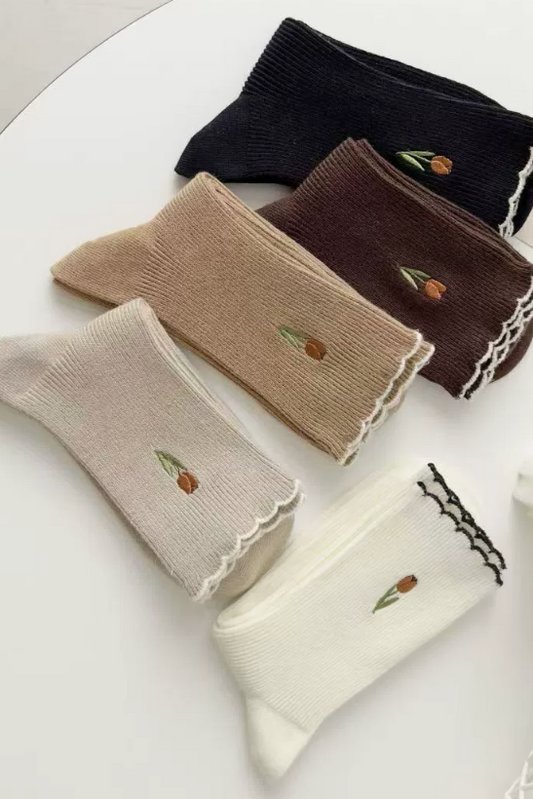 Four pairs of ribbed ankle socks arranged on a white surface, each with a small embroidered tulip near the top. The socks come in black, dark brown, taupe beige, and off-white/cream, and two have black lace trim while the others have white lace trim.