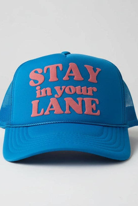 A bright blue trucker hat with pink embroidered text reading 'STAY in your LANE' on the front, shown against a light gray studio background.