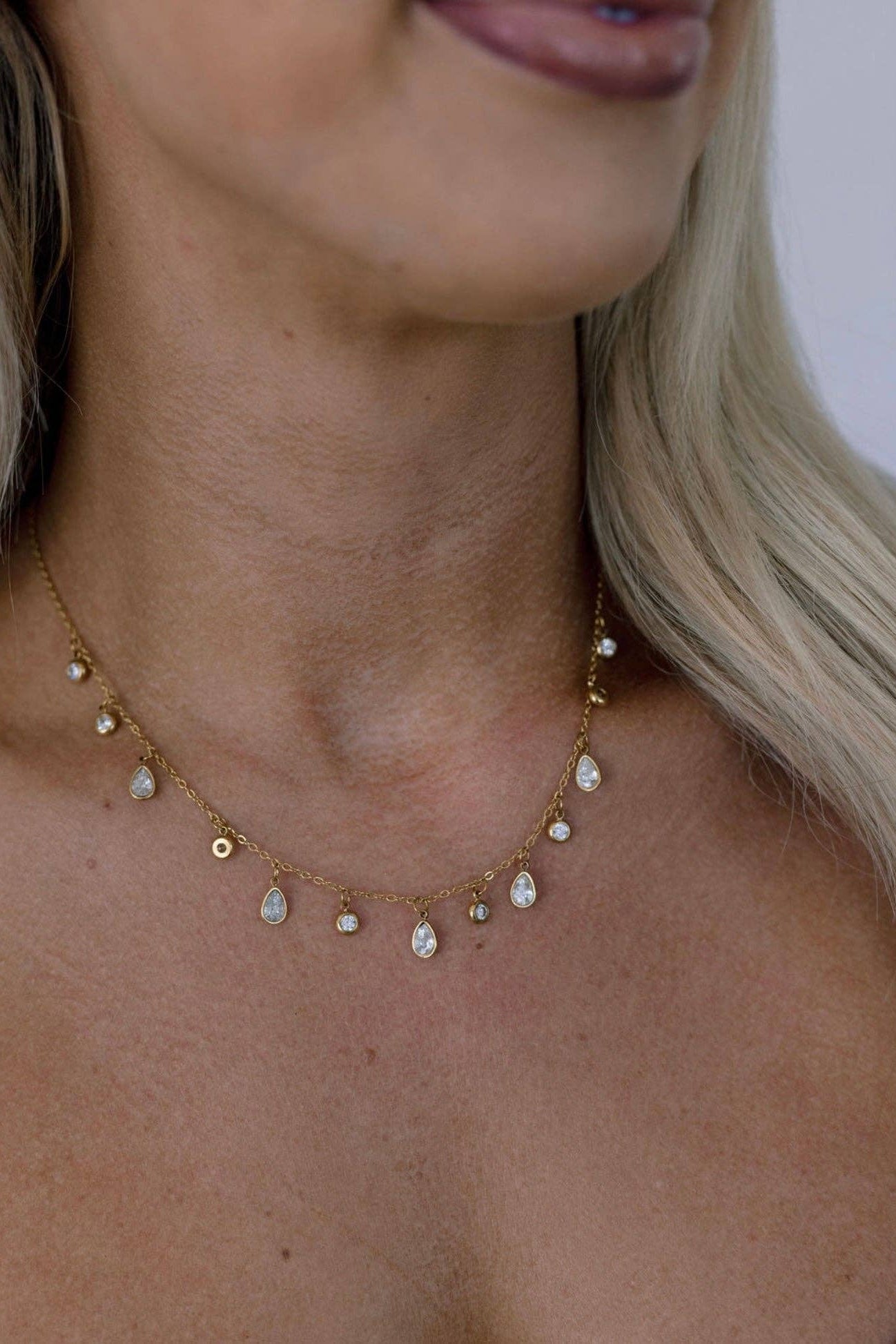 A close-up of a model wearing a delicate gold chain necklace adorned with small teardrop-shaped clear stones and tiny gold beads.