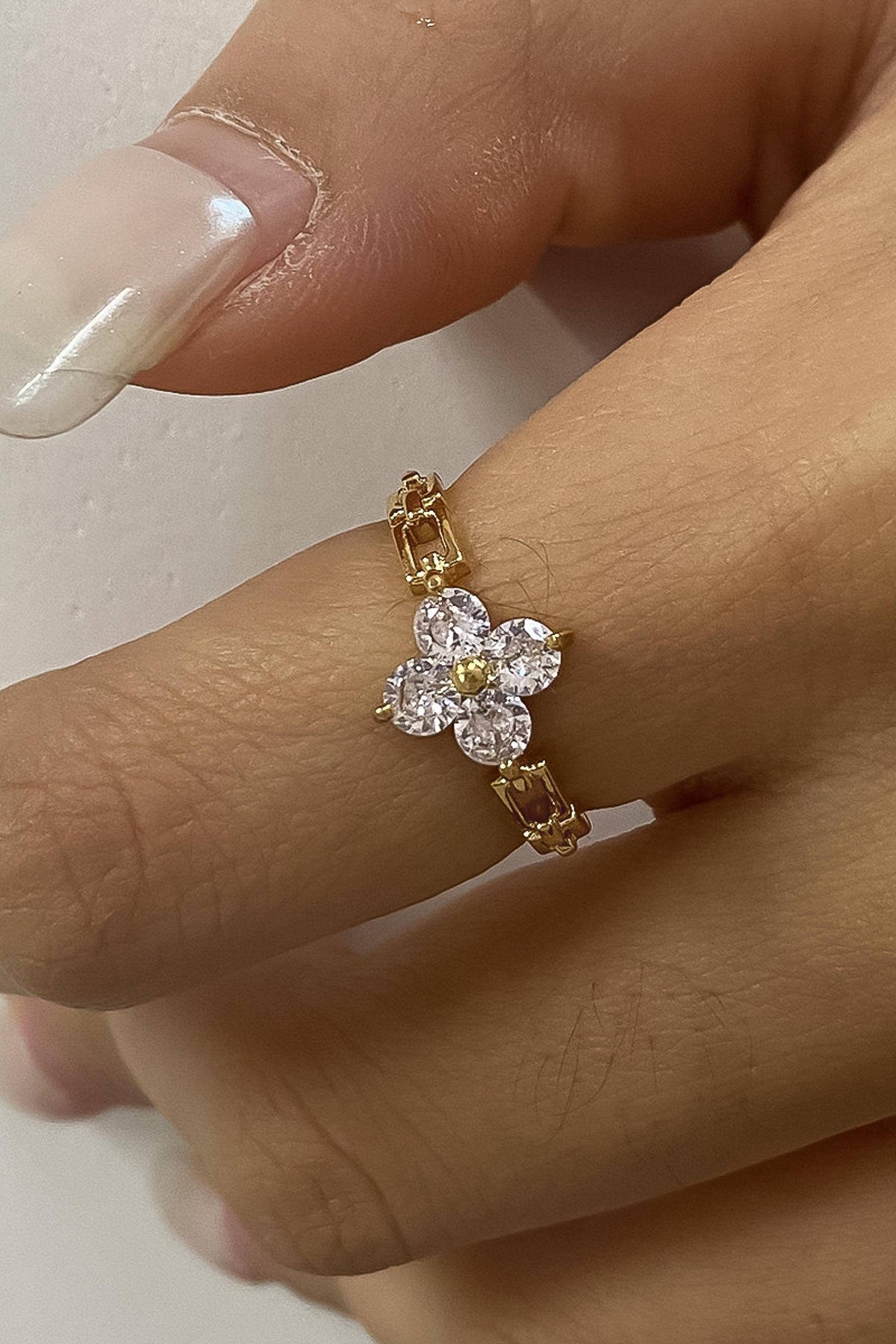 A gold ring featuring a flower-shaped cluster of clear gemstones, shown worn on a finger with a nude manicure.