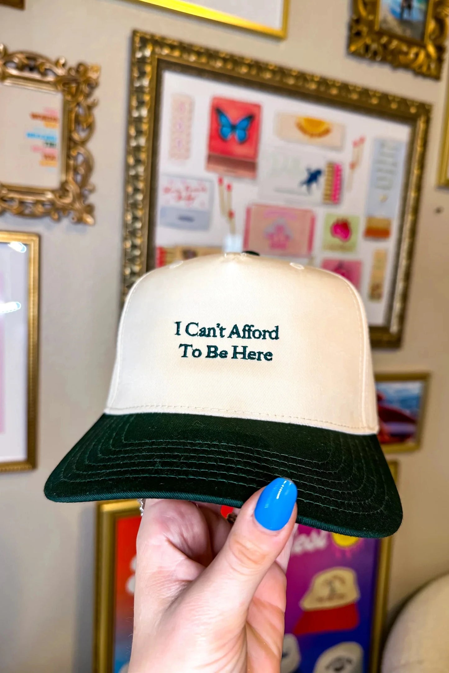 Trucker hat with 'I Can't Afford To Be Here' text held in front of a wall with framed pictures.