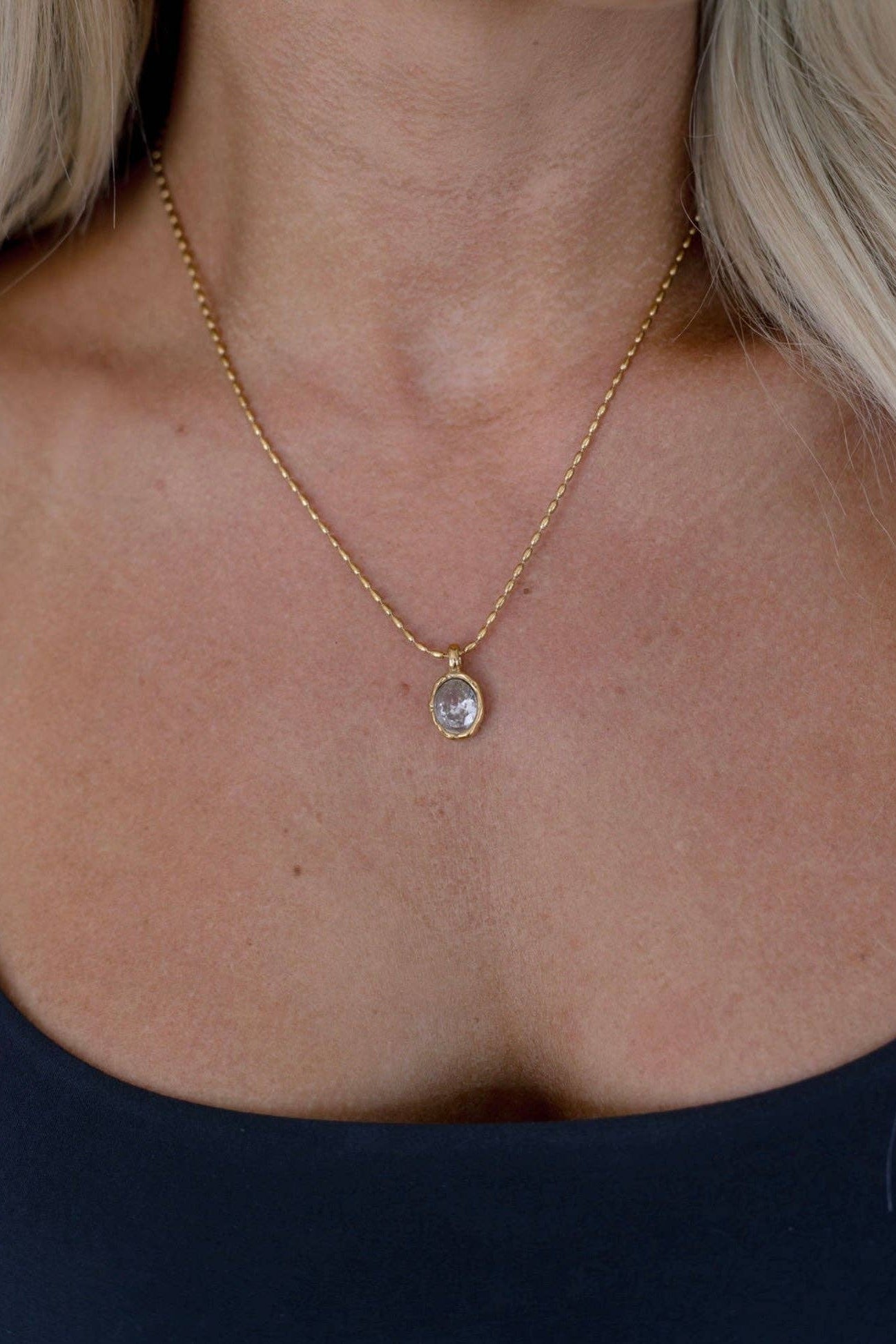 A close-up of a woman's neck and chest wearing a delicate gold chain necklace with a small oval clear stone pendant.