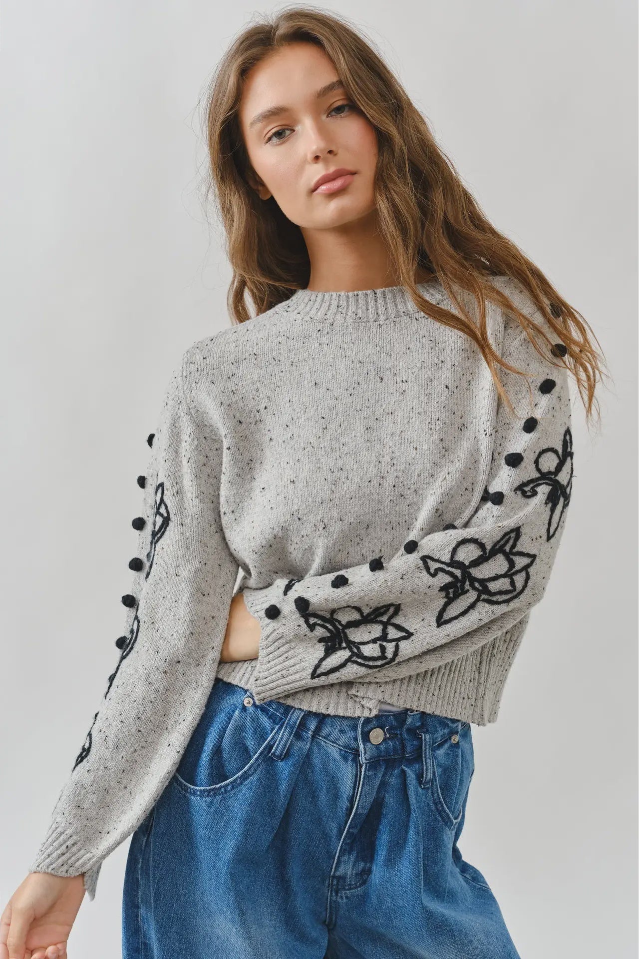 Woman wearing a gray sweater with black floral patterns and blue jeans on a light gray background