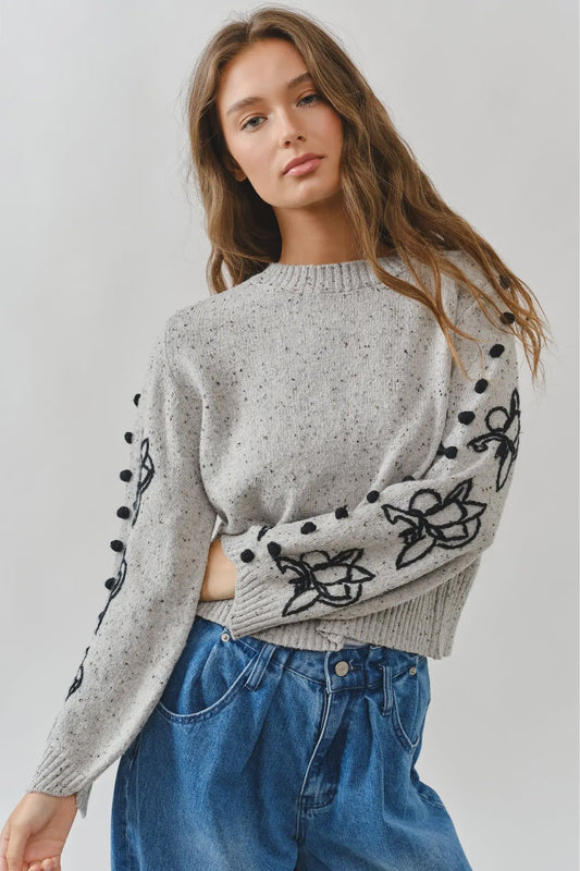 Woman wearing a gray sweater with black floral patterns and blue jeans on a light gray background