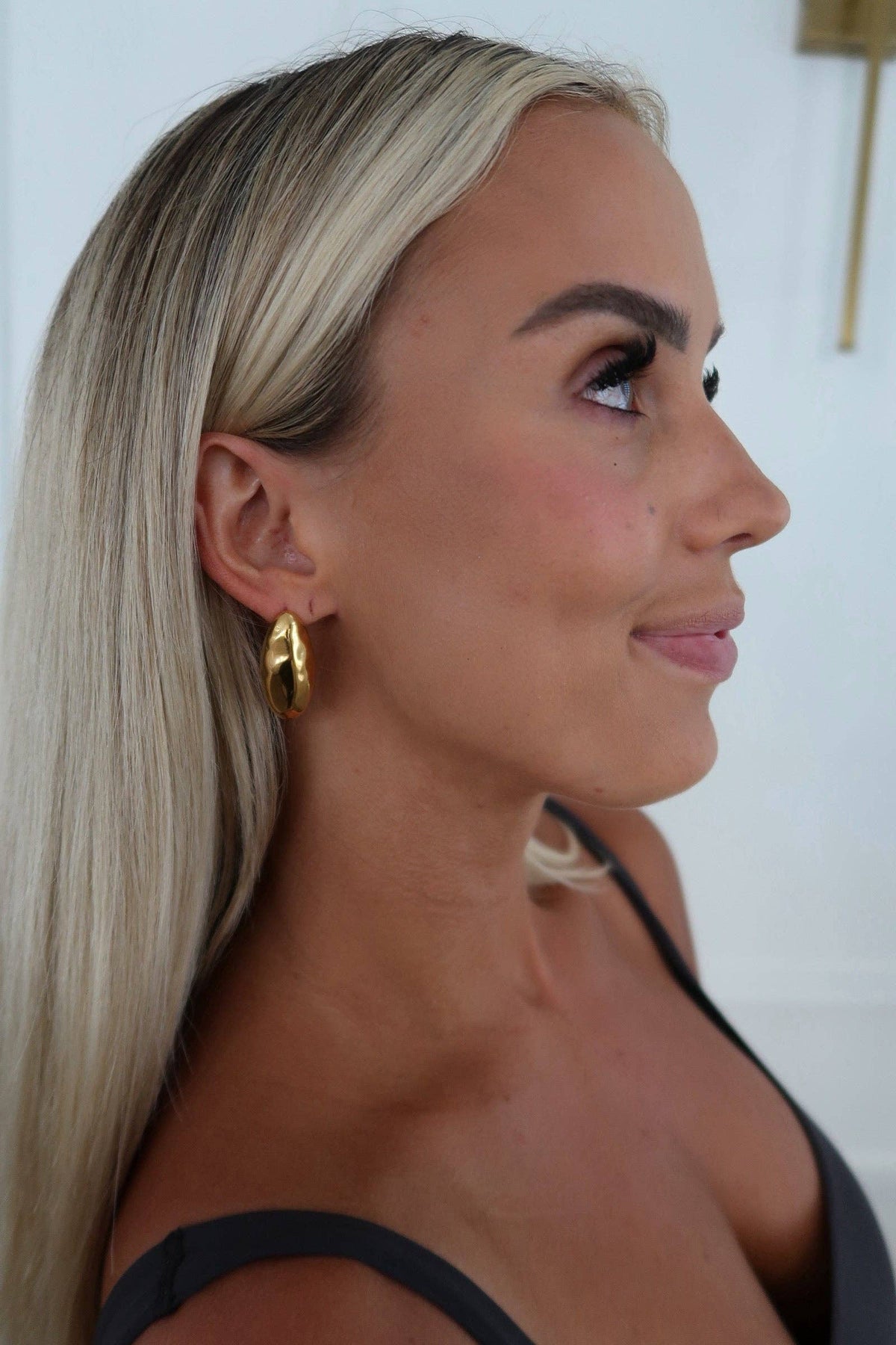 A close-up side profile of a woman with long blonde hair wearing a chunky gold hoop earring, set against a clean white background.