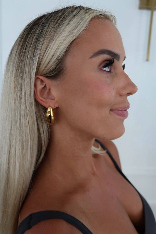 A close-up side profile of a woman with long blonde hair wearing a chunky gold hoop earring, set against a clean white background.