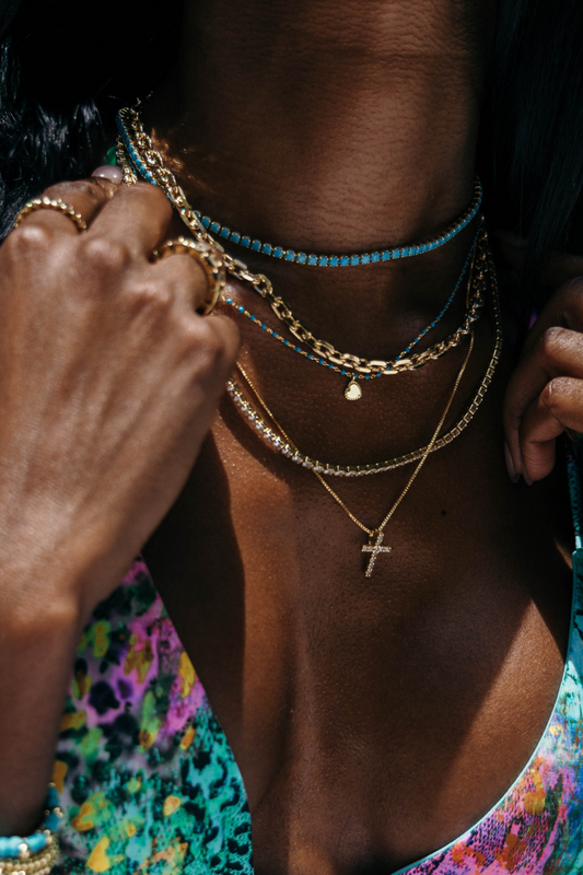 Person wearing multiple necklaces and bracelets with a colorful top, surrounded by greenery.
