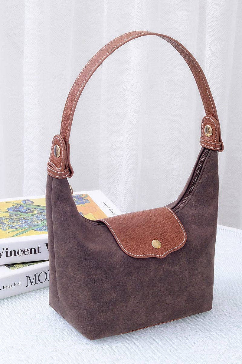 A brown suede hobo handbag with tan leather trim and a single shoulder strap, displayed on a white surface with a book and white curtain in the background.