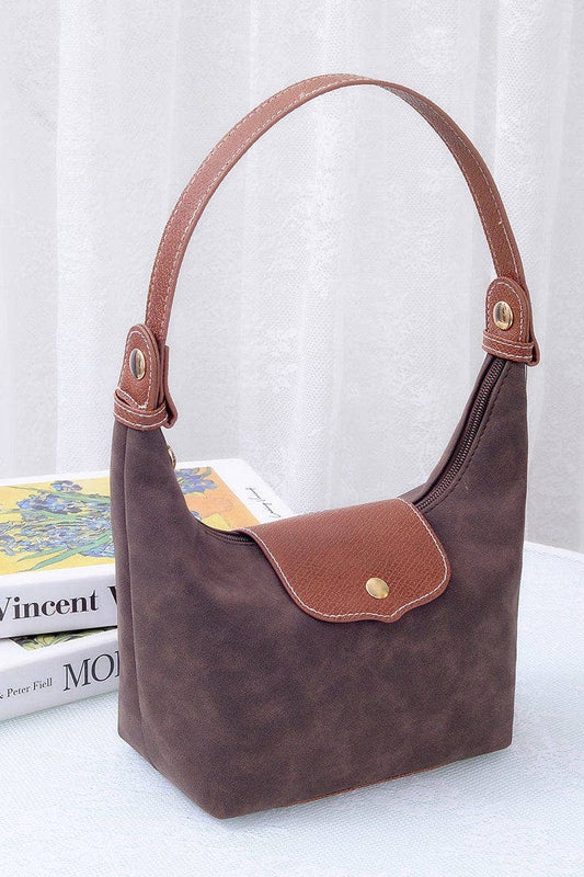 A brown suede hobo handbag with tan leather trim and a single shoulder strap, displayed on a white surface with a book and white curtain in the background.
