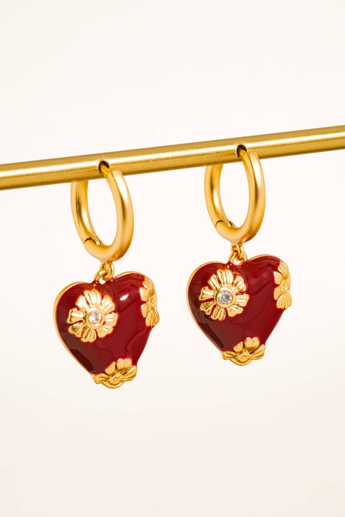 A pair of gold hoop earrings with heart-shaped red enamel pendants decorated with gold floral details and a small clear stone, hanging from a gold display bar against a light cream background.