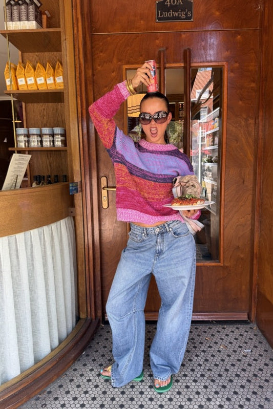 A woman posing in front of a wooden door wearing a colorful striped knit sweater with pink, purple, and orange tones, light blue wide-leg jeans, and green sandals, holding a plate of food and a drink can.