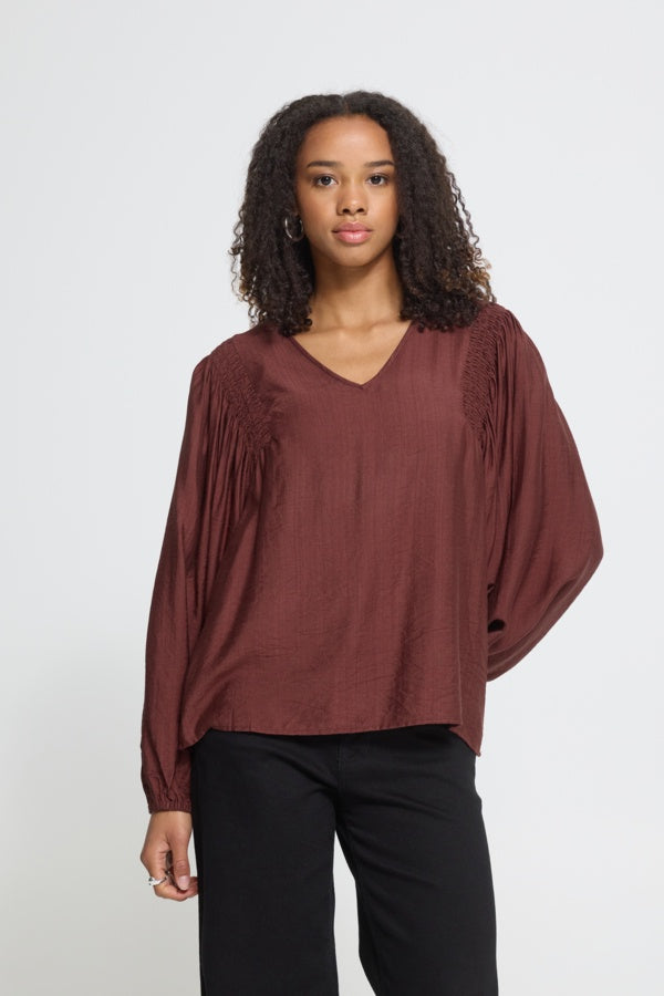 A woman modeling a rust-brown long-sleeve blouse with textured lace-up details on the shoulders, paired with black pants, against a light gray studio background.