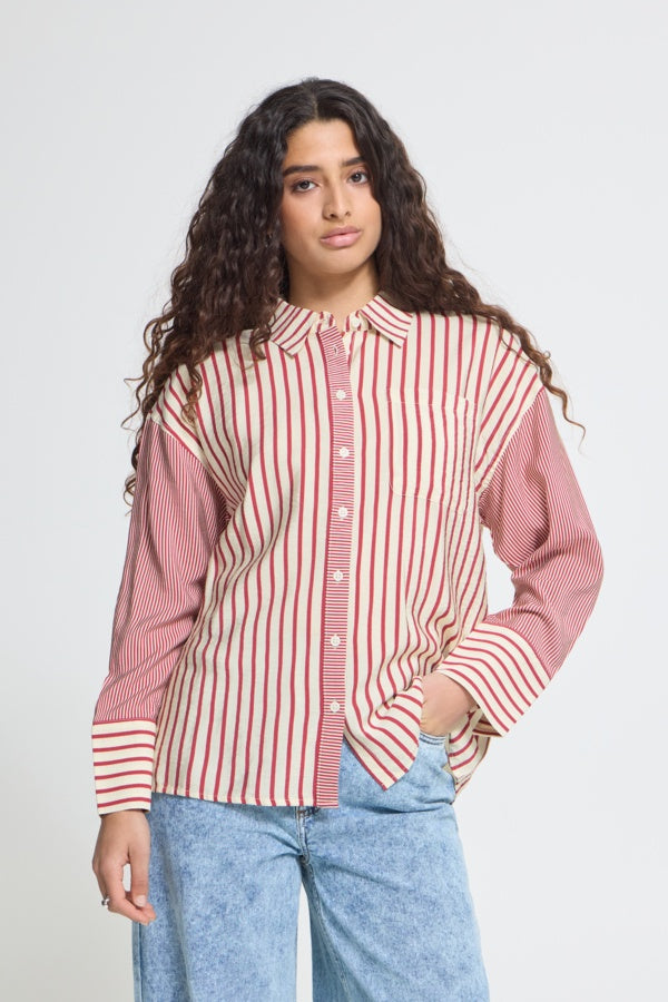A woman with long, curly dark hair models a pink and red vertically striped button-down shirt with light blue jeans against a plain light gray studio background.