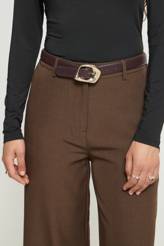 A close-up of a model wearing brown wide-leg trousers with a dark brown belt featuring a large gold-tone buckle, styled with a black long-sleeve top.