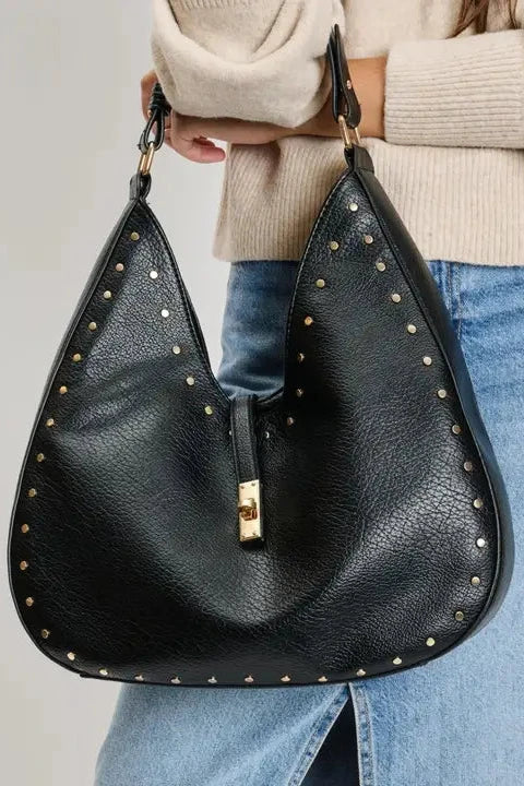 Black studded handbag held by a person wearing a beige sweater and blue jeans on a light gray background