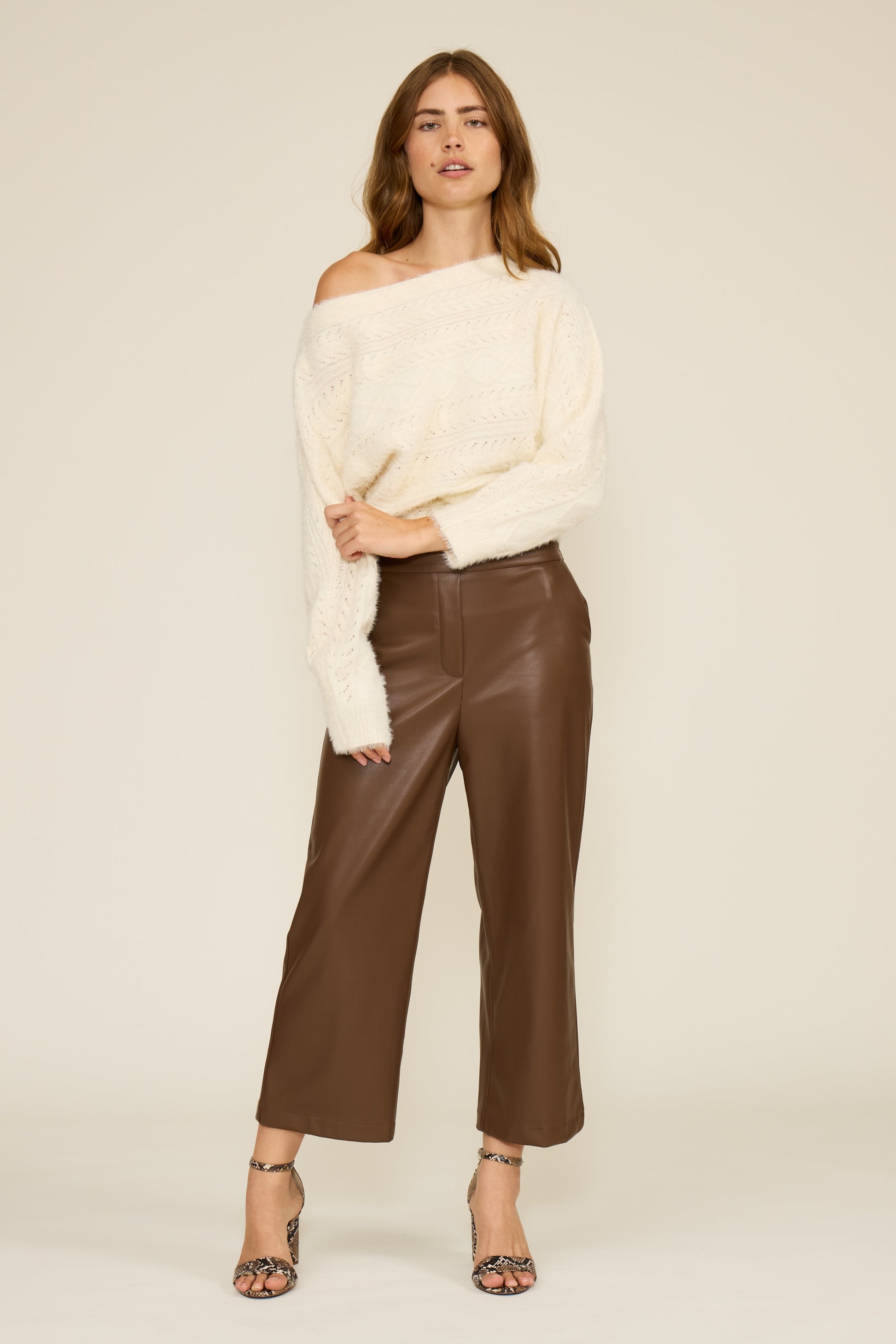 Woman wearing a white off-shoulder sweater and brown leather pants on a beige background