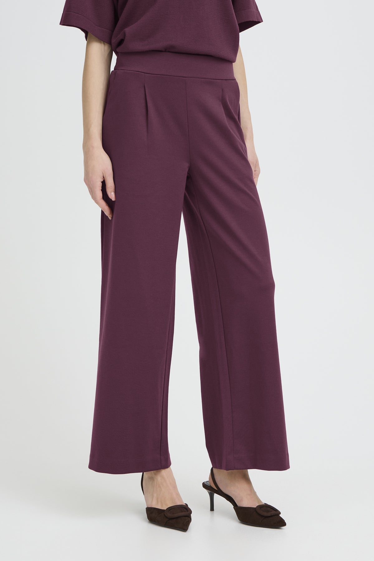 A model wearing a pair of high-waisted, wide-leg trousers in a deep plum purple color, paired with a matching top and dark brown pointed-toe heels, set against a plain light gray studio background.