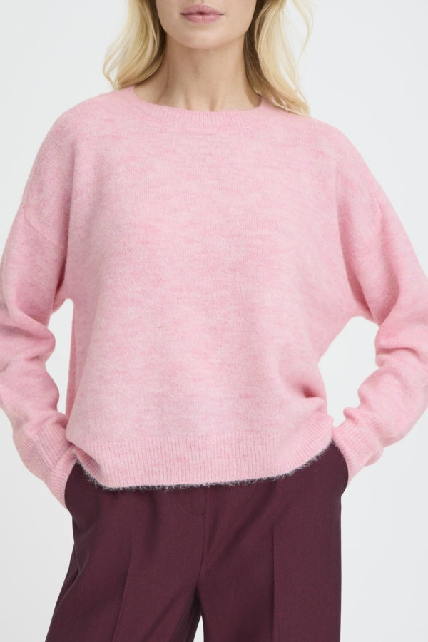 A woman wearing a light pink knit sweater with a crew neck and long sleeves, paired with dark burgundy trousers, shown from the chin down against a plain light background.