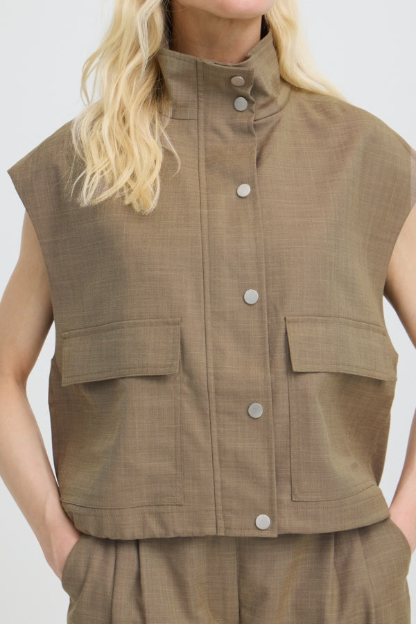 A close-up of a model wearing a sleeveless cropped vest in a textured taupe-brown fabric, featuring a high stand-up collar, silver-tone snap buttons down the front, and two large flap pockets on the chest.
