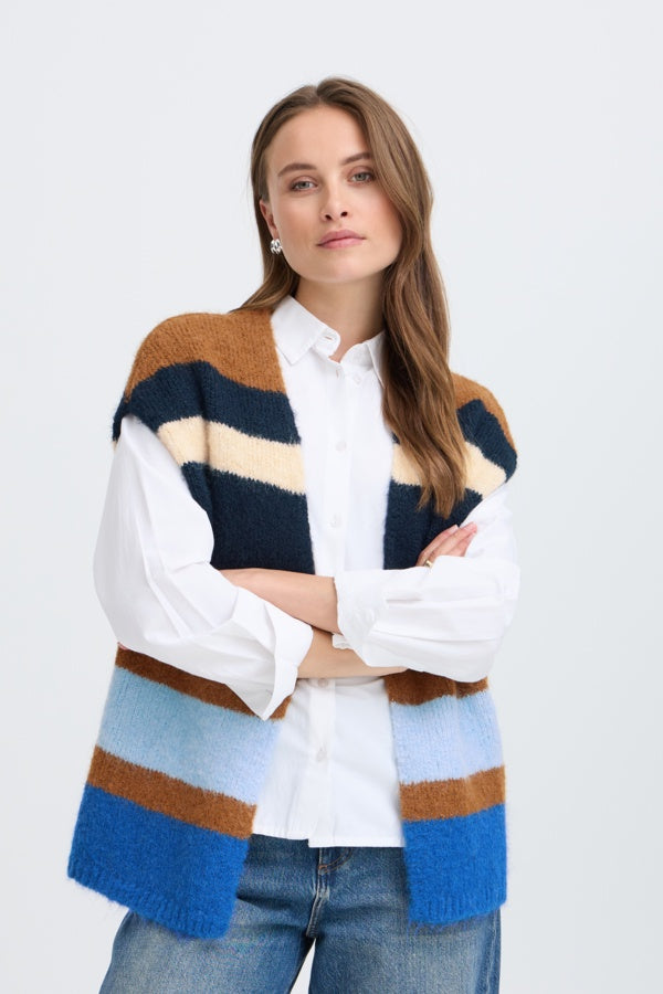 A woman wearing a fuzzy striped knit cardigan vest over a white button-up shirt and blue jeans, standing against a light gray studio background.