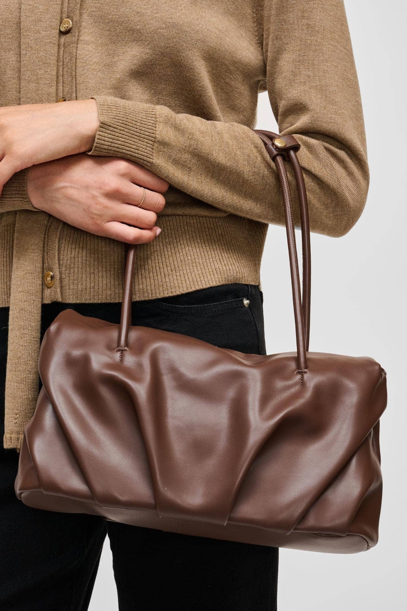 A person wearing a tan cardigan and black pants holds a brown slouchy leather handbag with two handles against a light gray background.