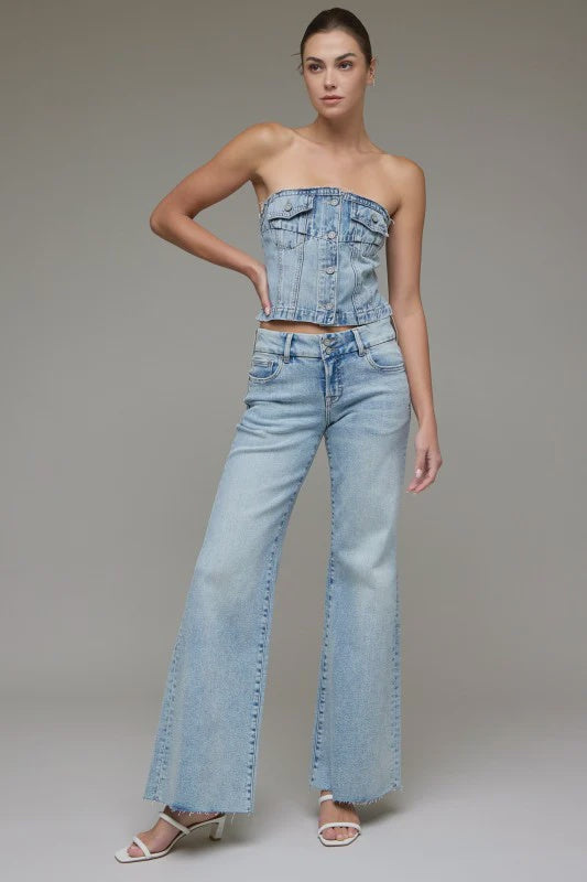 A model wearing a light-wash blue denim set consisting of a strapless cropped corset-style top and matching wide-leg jeans, paired with white heeled sandals, against a neutral gray studio background.