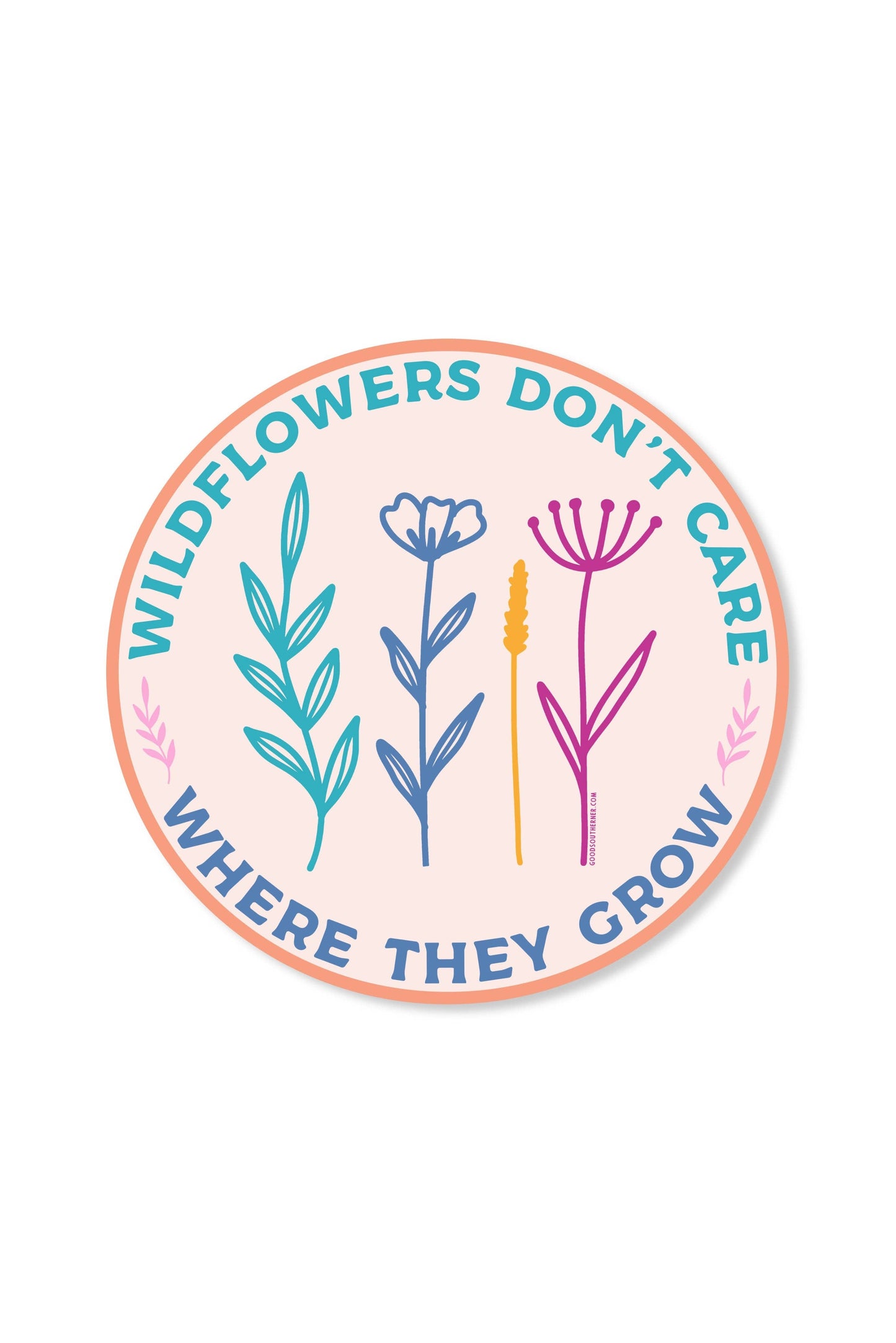 A round sticker or decal with a peach border and a pale pink center, featuring the text 'WILDFLOWERS DON'T CARE WHERE THEY CROW' in blue around three illustrated wildflower plants in teal, blue, yellow, and pink.