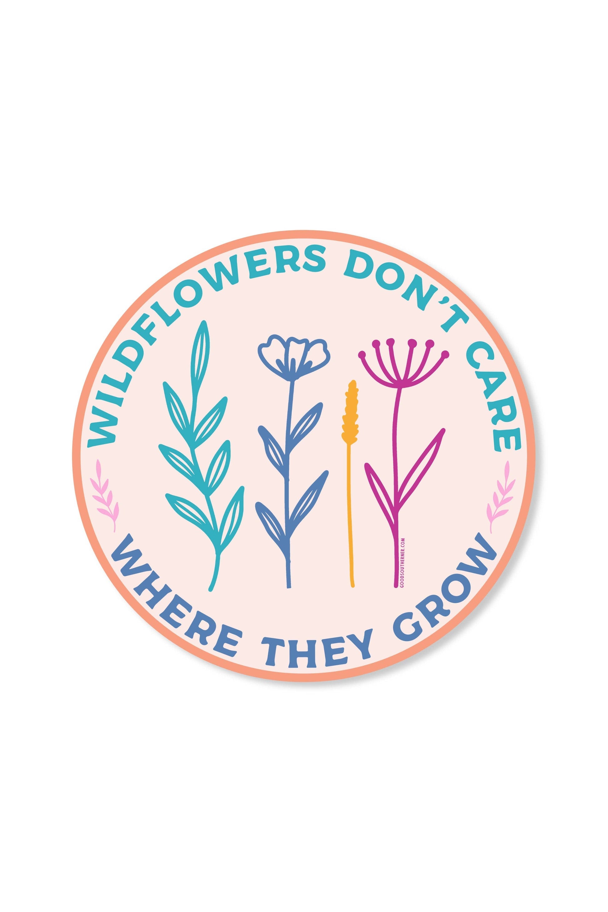 A round sticker or decal with a peach border and a pale pink center, featuring the text 'WILDFLOWERS DON'T CARE WHERE THEY CROW' in blue around three illustrated wildflower plants in teal, blue, yellow, and pink.