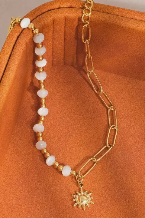 Gold chain bracelet with white beads and a sun charm on an orange fabric background