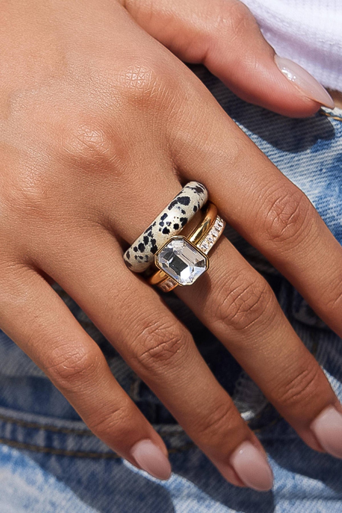 A close-up of a hand wearing three stacked rings: a gold band with a clear rectangular gemstone, a gold band with small clear stones, and a black-and-white spotted ring, set against blue denim.