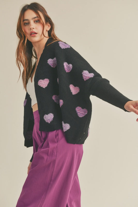 Woman wearing a black cardigan with pink heart patterns and purple pants on a beige background