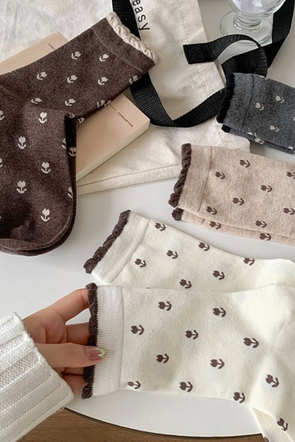 A hand wearing a cream-colored knit sock with a small brown floral pattern and dark brown ruffled trim, shown alongside other socks in brown, gray, and beige on a white surface with a book and bag as props.