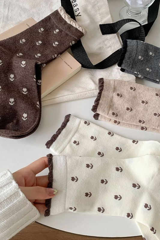 A hand wearing a cream-colored knit sock with a small brown floral pattern and dark brown ruffled trim, shown alongside other socks in brown, gray, and beige on a white surface with a book and bag as props.