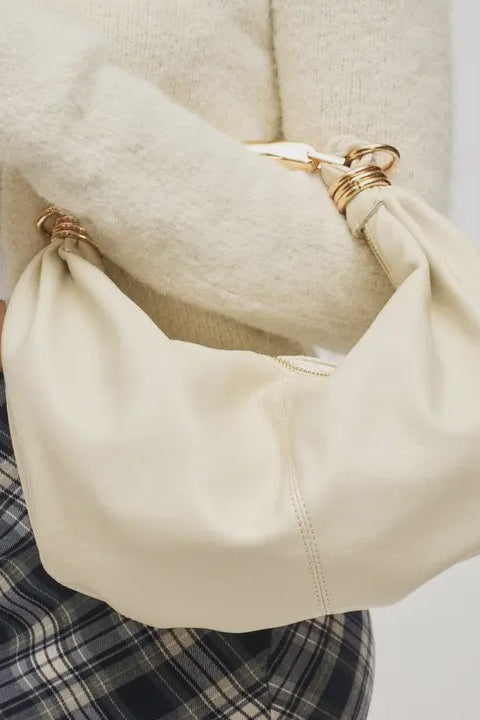 Beige handbag held by a person wearing a white sweater and plaid pants on a light gray background