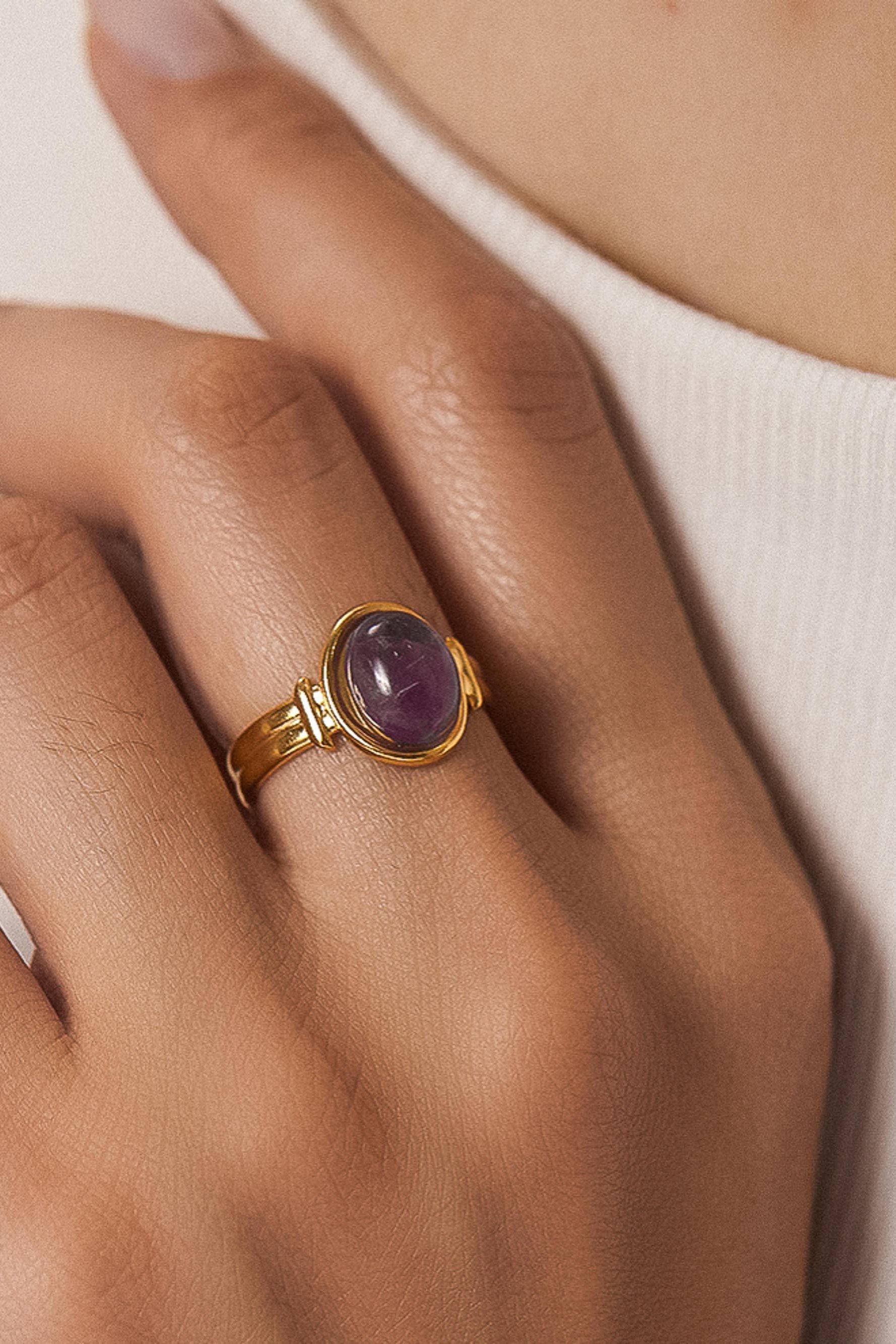 Gold ring with a purple gemstone on a hand against a neutral background