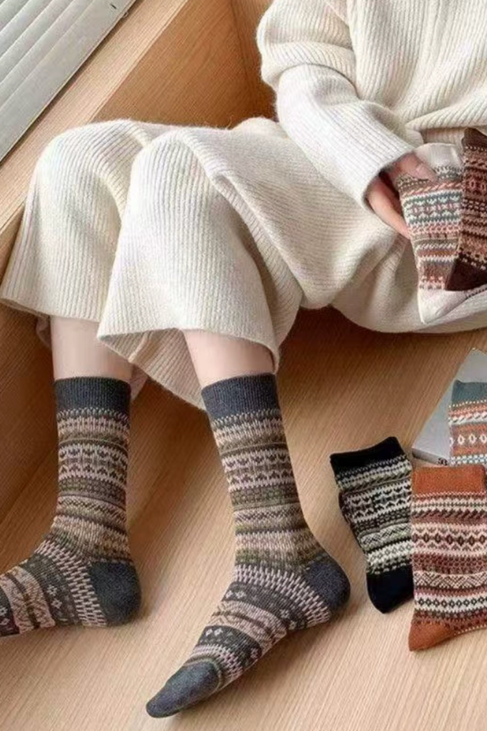 A person wearing a cream knit sweater and matching knit pants sits on a wooden floor, with patterned socks visible on the feet and additional socks placed beside them. The socks have a multicolor Fair Isle style design with dark gray, beige, brown, black, and rust tones.