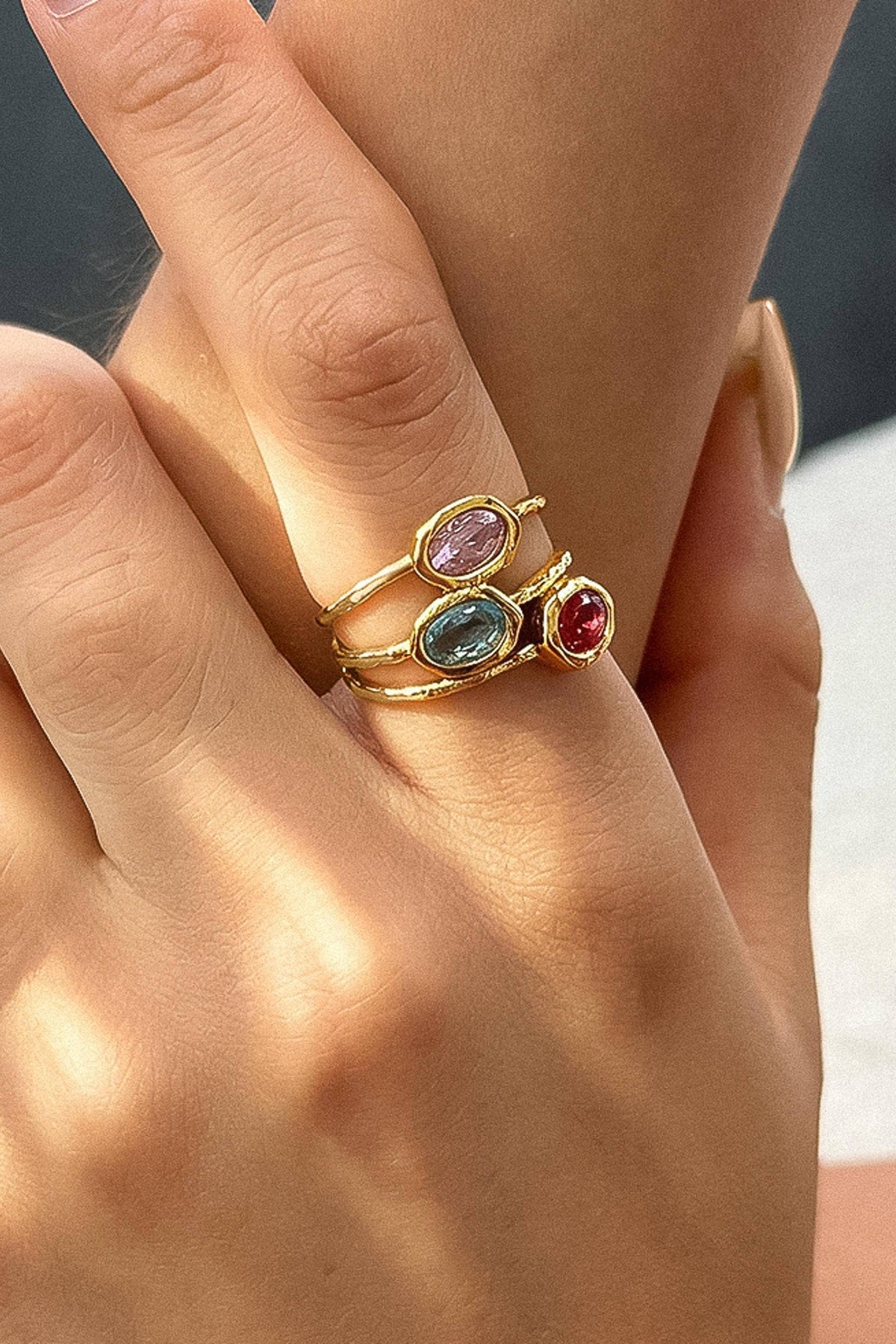 A close-up of a hand wearing a stack of three gold rings, each set with a different oval gemstone in pink, light blue, and red, resting on another hand.