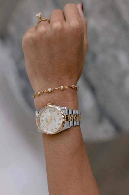 A close-up of a person's wrist wearing a two-tone gold and silver watch with a white dial, along with a delicate gold bracelet and ring, against a soft gray background.