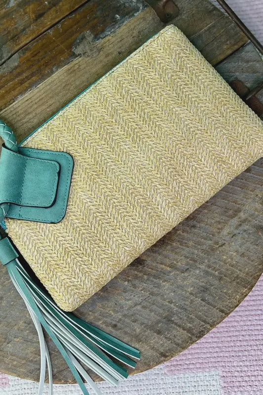 A beige straw wristlet with a green leather wrist strap and a tassel, displayed on a wooden surface.