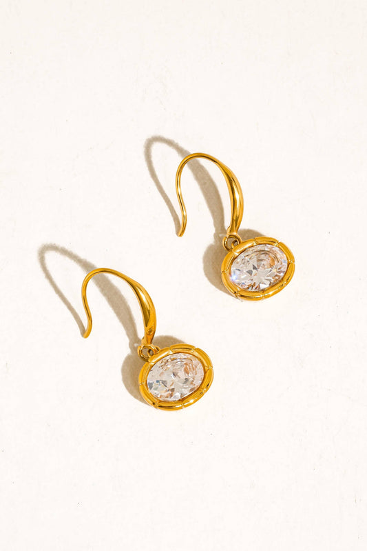 A pair of gold drop earrings with round clear faceted gemstones in bezel settings, displayed on a warm off-white background.
