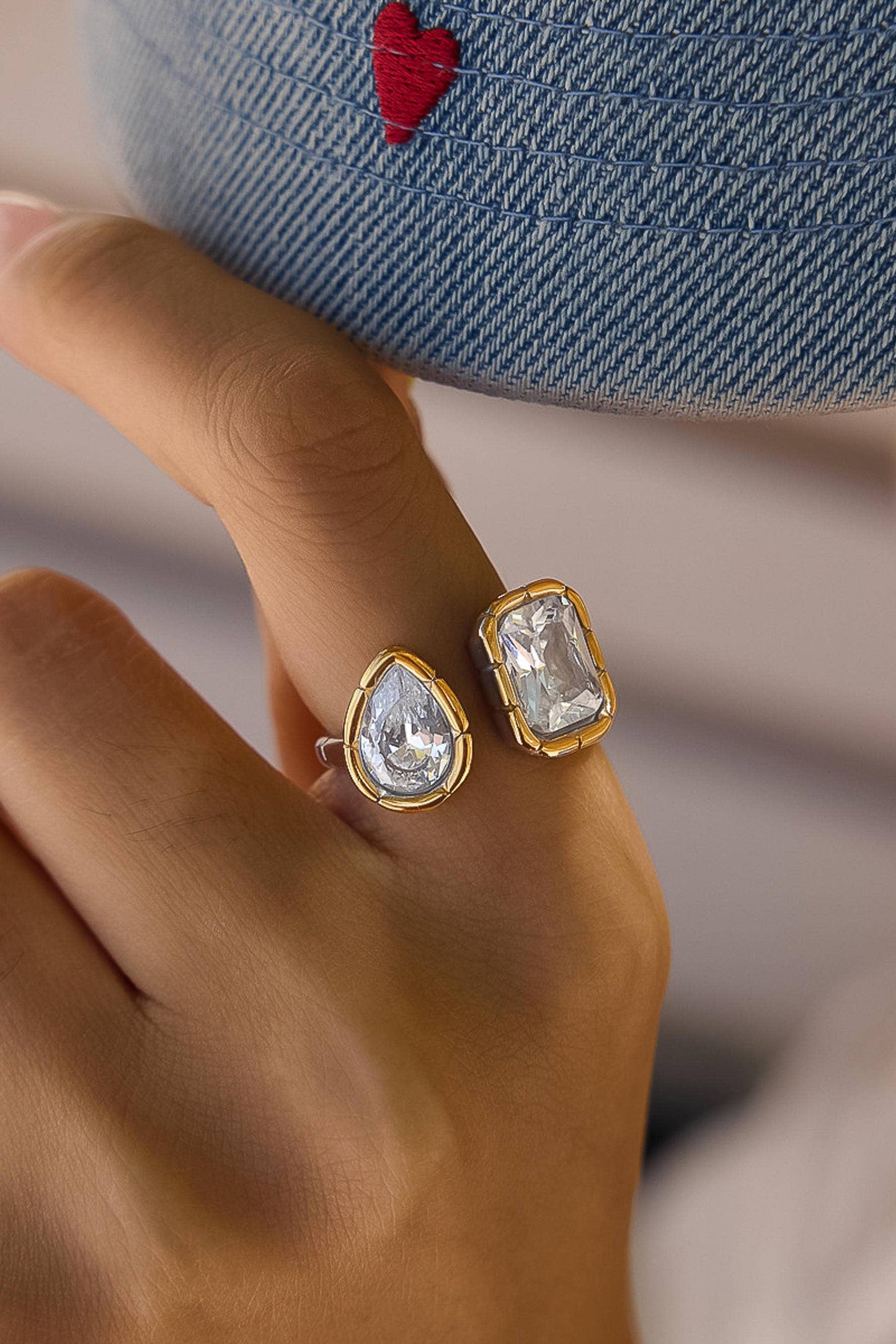 Two gold rings with clear teardrop and rectangular gemstones worn on a finger, shown in a close-up lifestyle shot with a denim cap in the background.