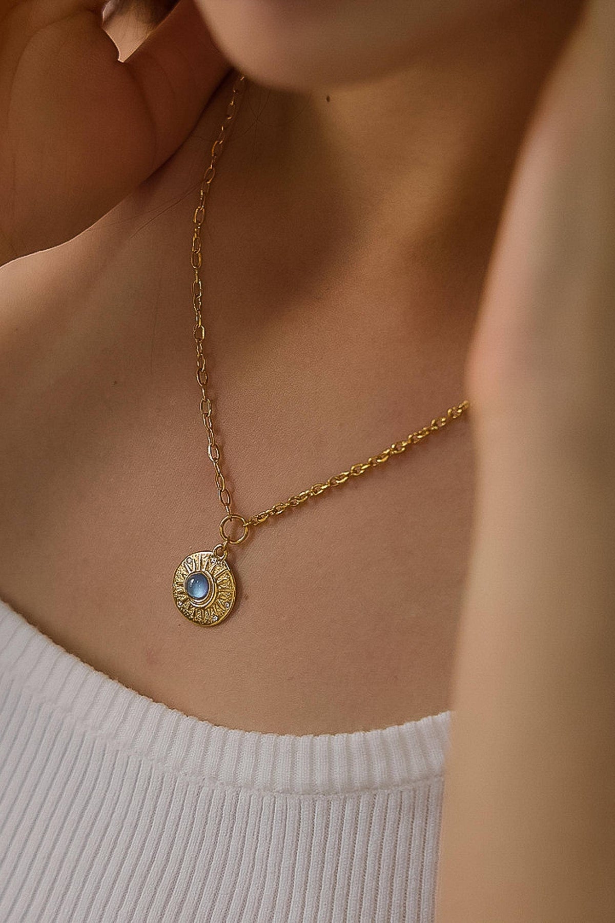 A close-up lifestyle shot of a model wearing a gold chain necklace with a circular sunburst pendant featuring a light blue center stone.