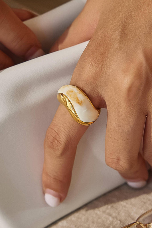 A close-up of a hand wearing a wide, sculptural ring with a polished gold-toned band and a large organic-shaped white stone with subtle warm marbling, resting on a white surface.