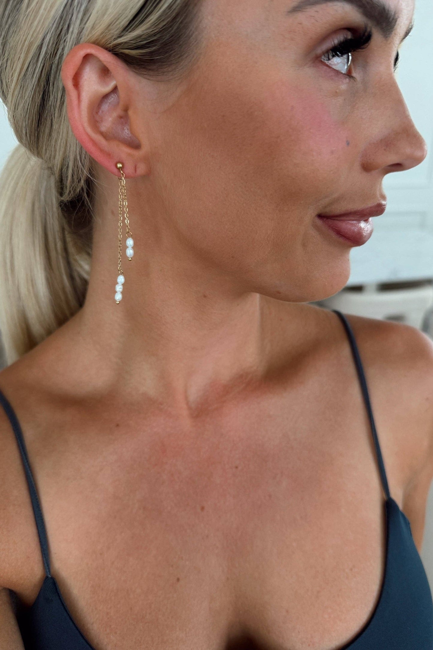A close-up side profile of a blonde woman wearing a delicate gold dangle earring with small white pearl-like beads, paired with a dark navy spaghetti-strap top.
