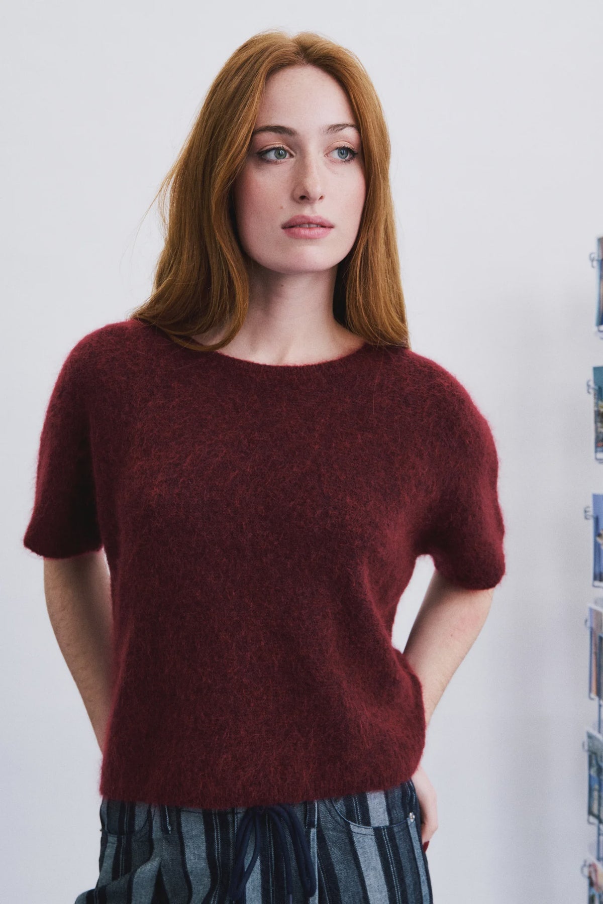 A woman with red hair wearing a short-sleeved, fuzzy burgundy knit sweater and striped trousers, standing against a light gray wall.
