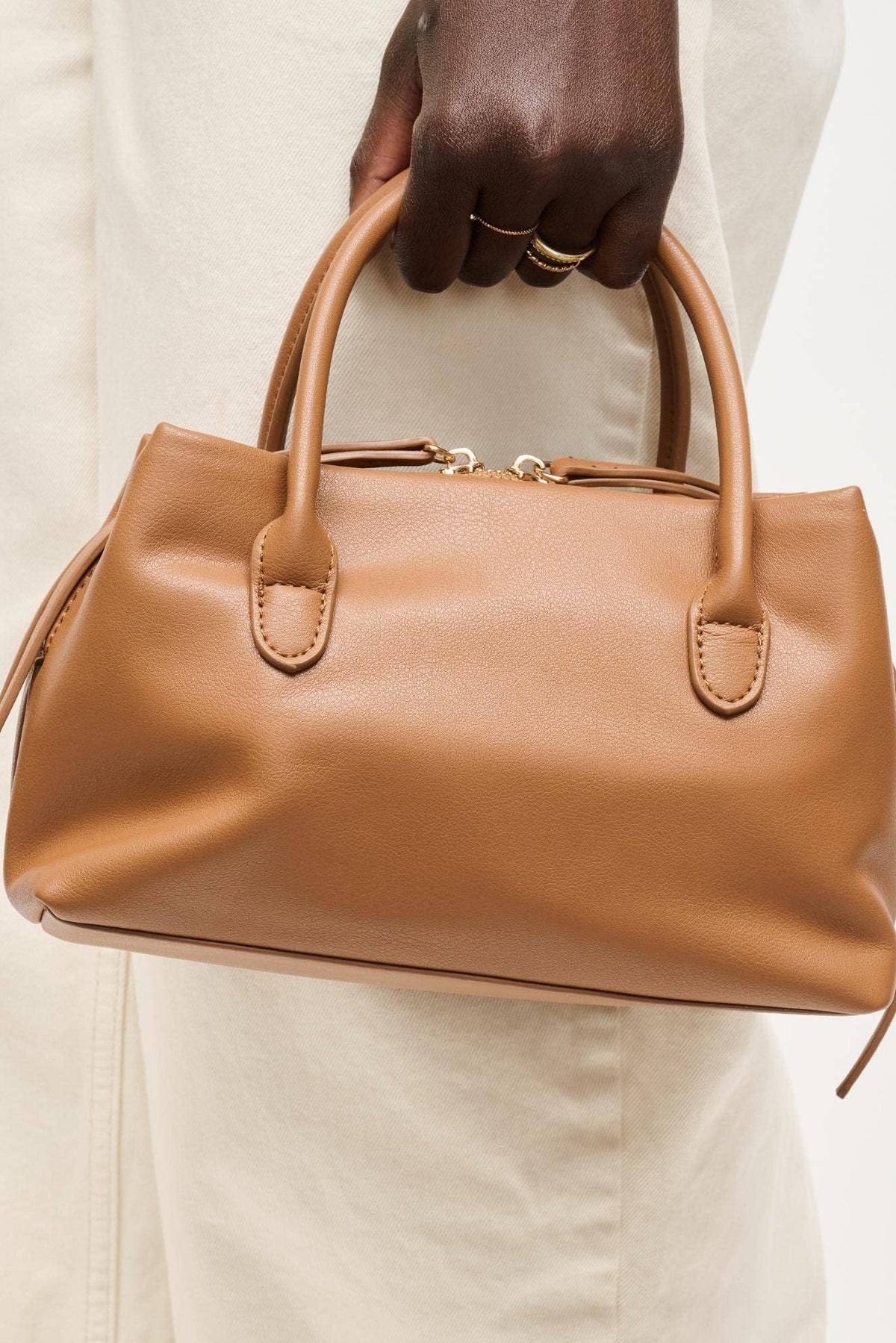 A tan leather handbag with double top handles and side ties, held by a person wearing a cream-colored dress against a plain white background.